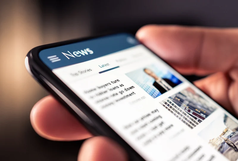 Image of a hand holding a smart phone showing articles in a news feed.
